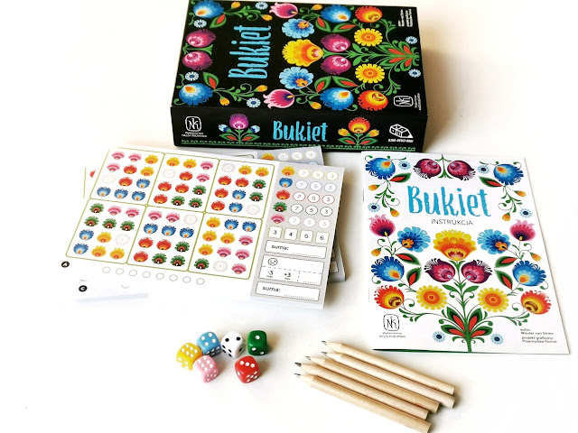 zdjęcie przedstawia pudełko od gry, dwa bloczki z planszami do gry, obok lezy instrukcja a przed nią pięć kostek w różnych kolorach i pięć ołówków