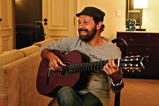  foto colorida de João Bosco sentado em casa tocando violão   