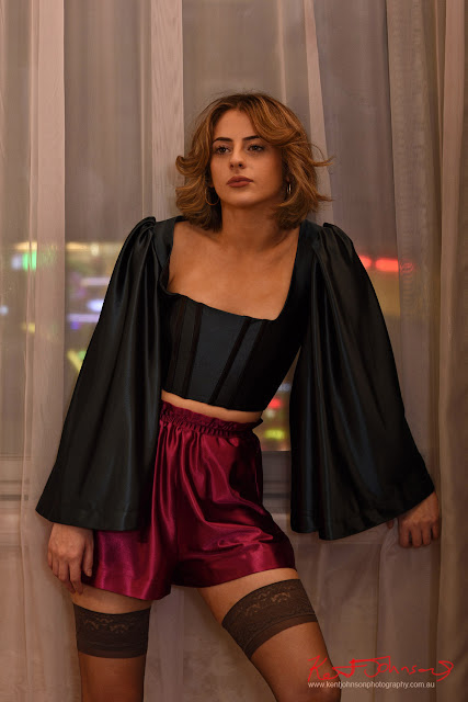 Midshot of a model standing before a tall window at night in bell sleeve, coreset style top with square neckline, soft red satin shorts and stockings.