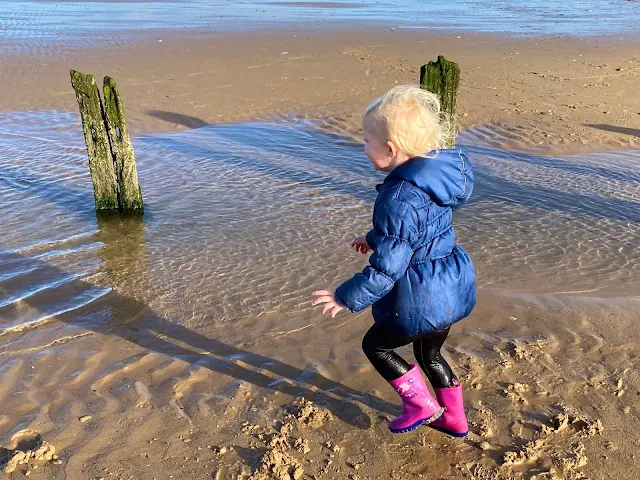 A 3 year old running around on the beach with very wet trousers and water sploshing out of the back of one of them