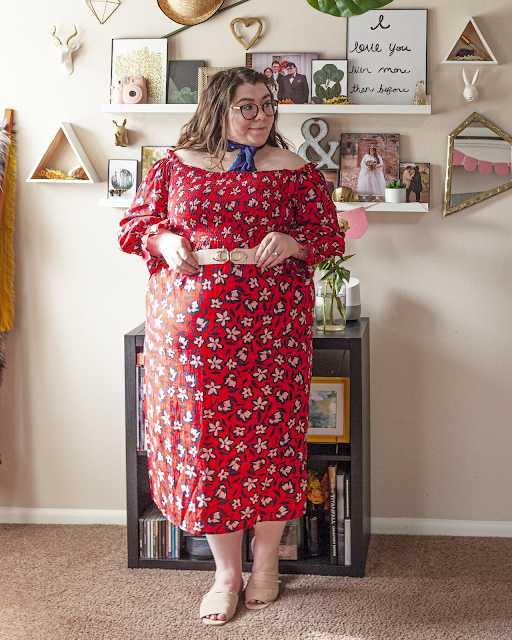 An outfit consisting of a navy blue square neck scarf tied around the neck, an off the shoulder red with navy blue outlined white flowers shirred bodice A-line midi dress with long bishop style sleeves, belted with a pink belt and pink slide sandals.