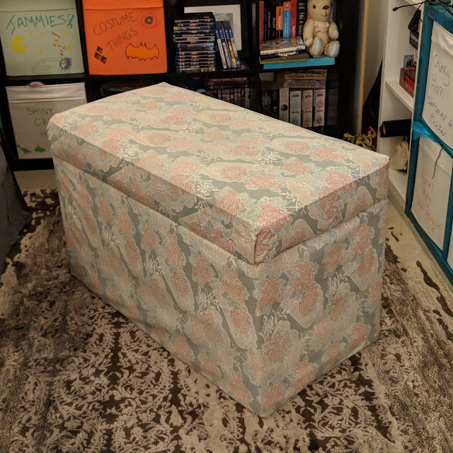 A photo of a loungeroom lined by shelving, the forefront of the photo is an ottoman covered in very faded fabric, which would have once been pink flowers on a blue background. The fabric is dirty and evidently a home job.