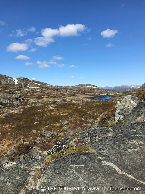 A dark blue lake on a flat high-altitude plateau covered in moss and grass, and in places with snow, under a light blue sky with a few fluffy white clouds.