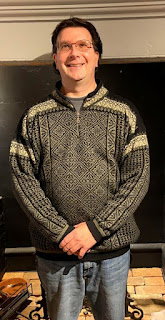 White man with dark hair and glasses wearing a sweater with ornate black and white pattern