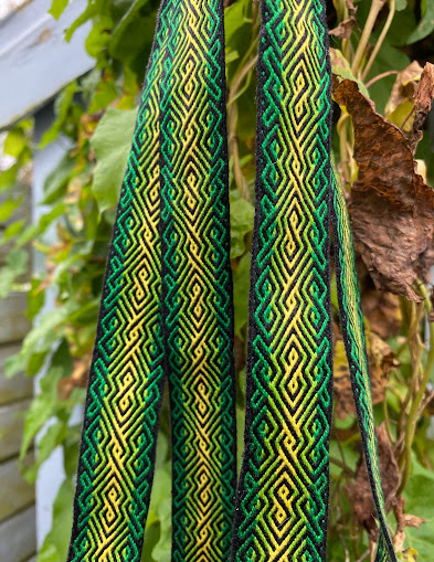 A tablet woven band decorated with yellow and green diagonal lines and infinity symbols on a black background hangs in front of leaves beginning to turn autumnal brown