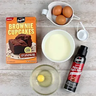 Ingredients for Brownie Cupcakes, including brownie mix, eggs, and nonstick spray, arranged on a wooden table.