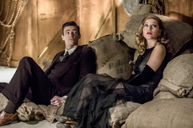 A screencap of Barry Allen and Kara Danvers sitting on a pile of flour sacks in a warehouse. They both wear 1920s clothing: Barry, a dark three-piece suit, and Kara, a black full length evening gown with a glittery bodice and elbow-length gloves.