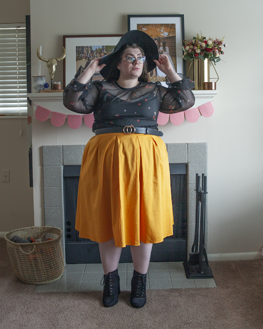 An outfit consisting of a black wide brim black hat, black sheer long sleeve microfloral dress as a top tucked into a yellow midi skirt and black lace up heeled boots.