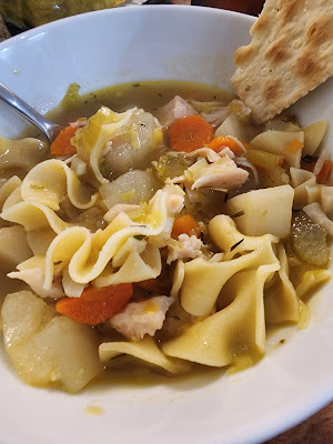 A heavily populated bowl of homestyle chicken noodle soup with saltine crackers. The bowl is white and filled with a thick stew. Chicken chunks, egg noodles, sliced carrots, diced potatoes, and sliced leaks sit in a glistening broth.