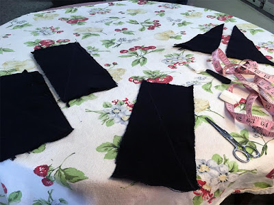 A few stacks of rectangular pieces of blue-black fabric, a small pair of scissors, an unspooled pink tape measure, and a pair of tall, skinny right triangles on blue-black fabric on a white floral tablecloth.