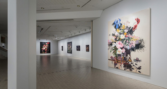 Kuvassa näkyy osa taidemuseo Poikilon alakerran näyttelytilasta. Seinille on ripustettu kukka-aiheisia maalauksia ja valokuvia.