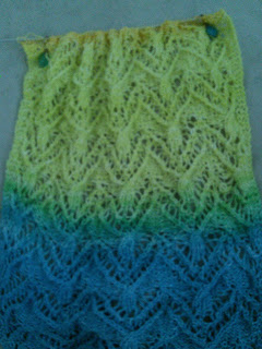 lace knitting with fingering-weight yarn done on a straight needle.  The yarn is a gradient blue-green-yellow, with orange just appearing on the needle.