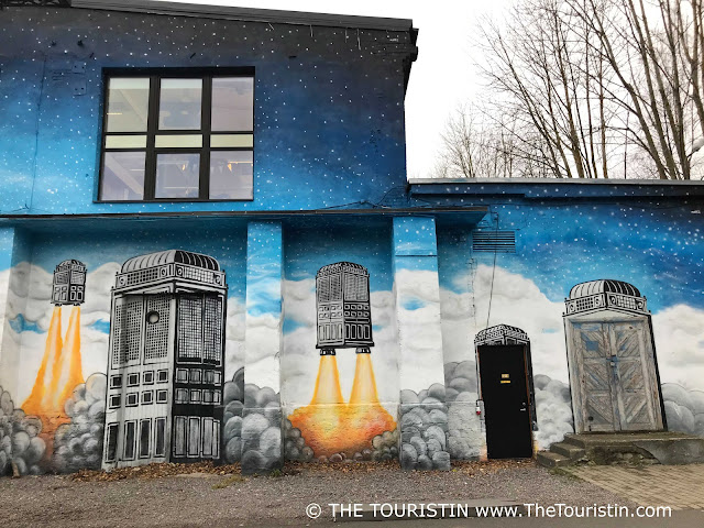 A mural with five black and white ufos, two of them start their journey into the sky, leaving thick grey clouds of smoke behind - a light-blue-sprinkled-with stars-washed brick facade as background.