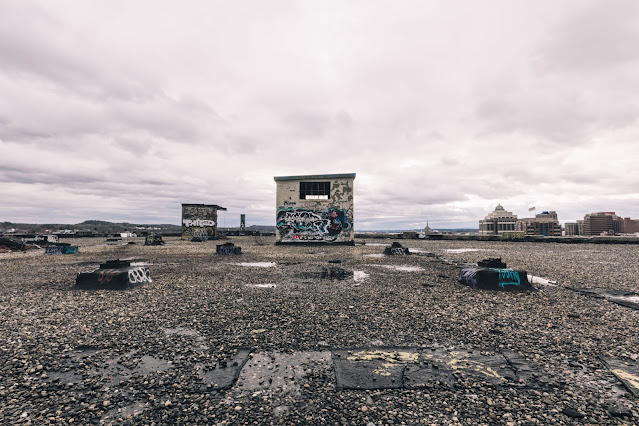 The flat rooftop of an urban building, dotted with graffiti-covered structures and debris under a brooding sky. Puddles reflect the cloudy heavens, while in the distance, the city skyline looms, contrasting the edgy foreground with its orderly array of buildings.