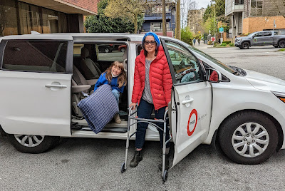 a woman stands with a walker in front of a Modo minivan with a girl sitting in the back seat. the sliding door & side door of the van are open, it's parked in a hospital parking lot