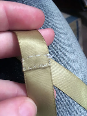 A close-up of sage green ribbon with the ends stitched together with a rectangle of tan thread, held between the fingers of a white hand.