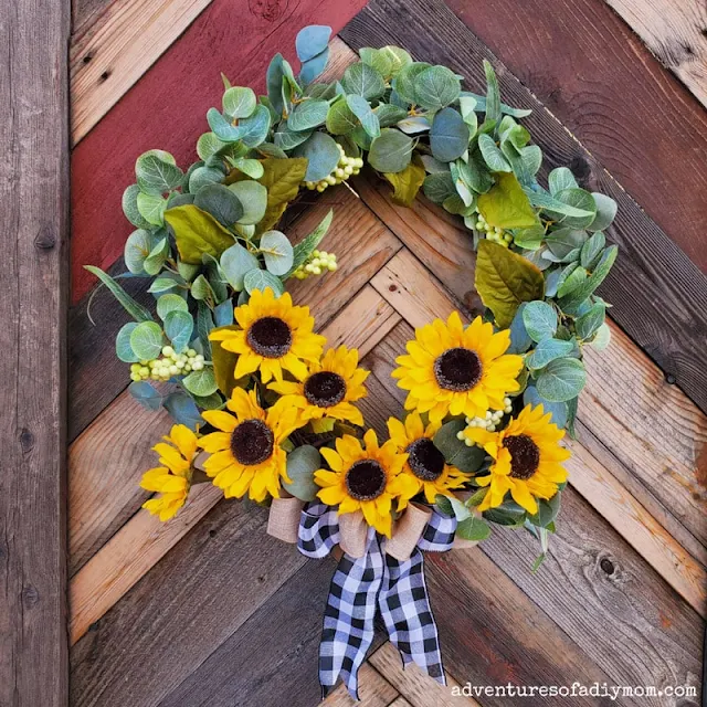 Sunflower wreath with lush greenery and buffalo plaid/burlap layered bow hanging on a rustic chevron fence panel outside.