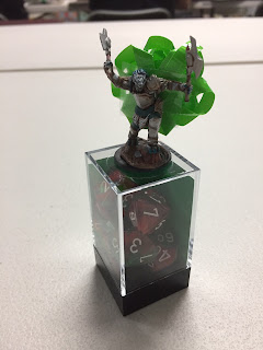 The Sadun miniature with a bow stuck to her back stands on a clear plastic dice box