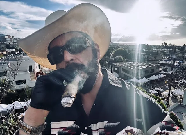 Sporting dark sunglasses, cream-colored cowboy hat sucking on a half-smoked stogie wearing leather gloves