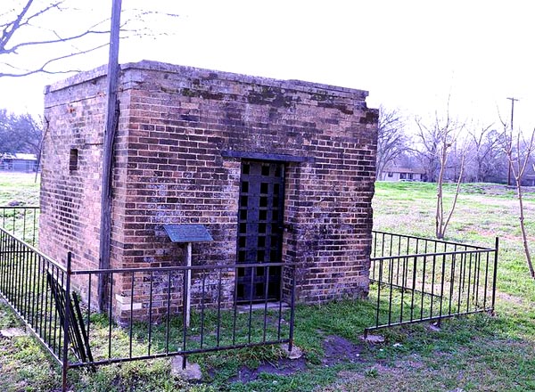 a small, square red-brick building with strapped iron doors stands alone in a grassy field, with a low wrought-iron fence surrounding it