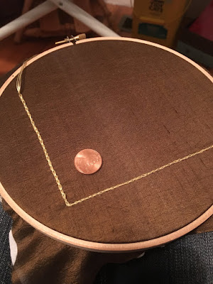 A right-angle formed of two gold chain stitch lines on olivey-brown silk in a wooden embroidery hoop. The right line is half the thickness of the left, and a penny is sitting in the corner they form.