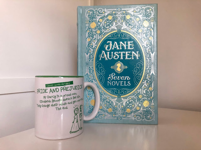 Pale blue and gold large novel standing up on white shelf reading 'Jane Austen 7 Novels' next to green and white pride and prejudice mug with illustrations of unhappy man and woman