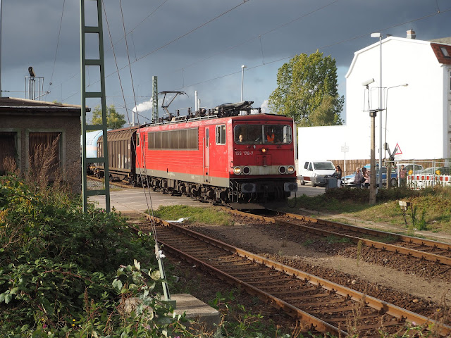 Über den Bahnübergang für eine eckige rote E-Lok der Bahn. Ein Güterwaggons istzu sehen. Rechts ein weißes Haus, links Teile eines kleinen beigen Hauses. Dunkle Wolken.