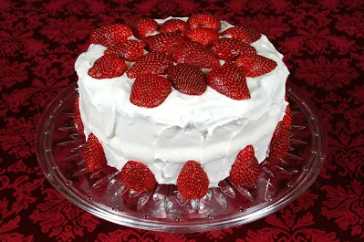 Auf dem Bild ist eine runde, weiße Torte zu sehen. Sie mit Erdbeeren garniert. Die Creme ist Sahne und wurde ungleichmäßig aufgetragen. Sie steht auf einem großen Glasteller und rund um liegen noch weitere Erdbeeren.