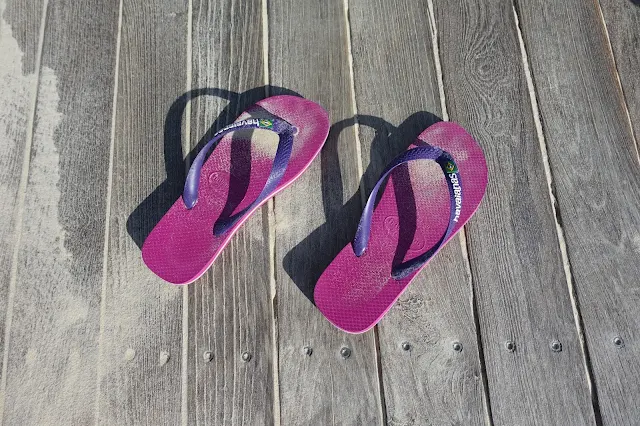 Pink and purple havaianas covered with sand on a wooden boardwalk dusted with sand