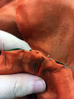 A close-up of a white finger and thumb holding the pinned edges of sheer peach and green-gold silks, with their edges turned in. The peach silk is a little bubbled between the pins.