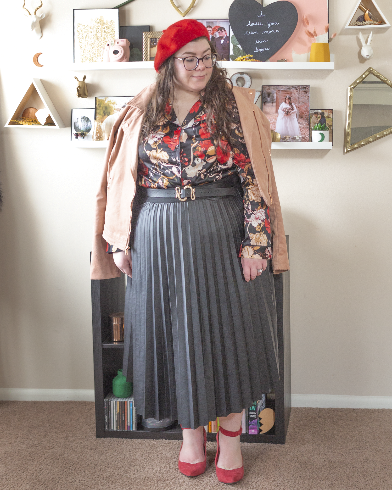 An outfit consisting of a red beret, a pink suede moto jacket draped over the shoulders of a black with cream, red and pink florals pajama style top tucked into a black pleated faux leather midi skirt and red ankle strap heels.