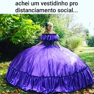 Foto em dia ensolarado. Em um recanto arborizado, gramado aparado com algumas folhas amareladas caídas. Uma mulher jovem, cabelos loiros compridos penteados e presos à esquerda por uma flor preta. Ela usa um vestido longo, roxo acetinado e super rodado (anágua de armação). A parte superior em tomara que caia, com franzido largo do mesmo tecido acompanha ao redor do decote, e cobre parte do corpete preto bem acinturado de onde incia a saia em circunferência de diâmetro exagerado. A saia finaliza com um recorte bem franzido e na costura do corte, um acabamento em renda preta.  No topo lê-se: achei um vestidinho pro distanciamento social...