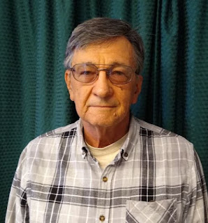 White man with slightly graying hair and glasses. He is wearing a plaid collared shirt with shades of white, gray, and black and standing in front of a dark green curtain.