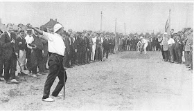 Black and white photo of a man in white polo shirt and black pants throwing a cricket ball while other players in white clothing stand to hit the ball. The cricket players are surrounded by men in various clothing, some wearing military uniform and others wearing suits.