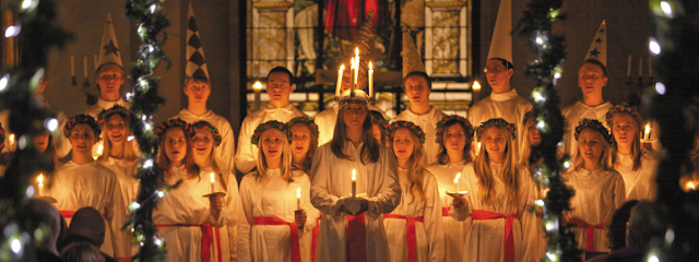 Young women in white robes with red sashes, hiolding candles celebrating the Scandivan tradtion of St Lucia's Day