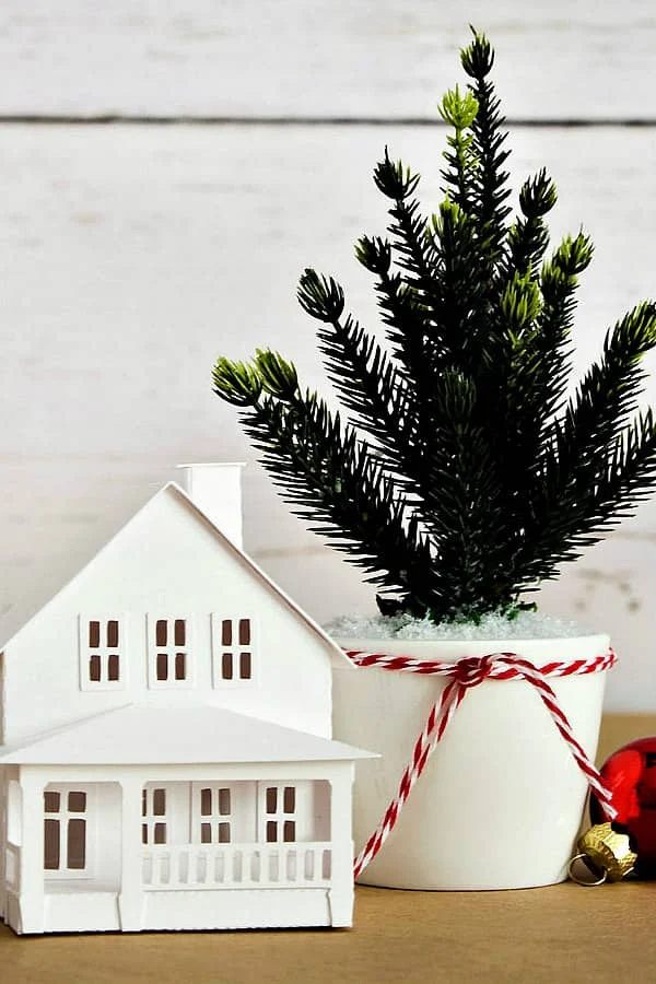 tiny white paper house on table next to potted evergreen with Christmas baubles