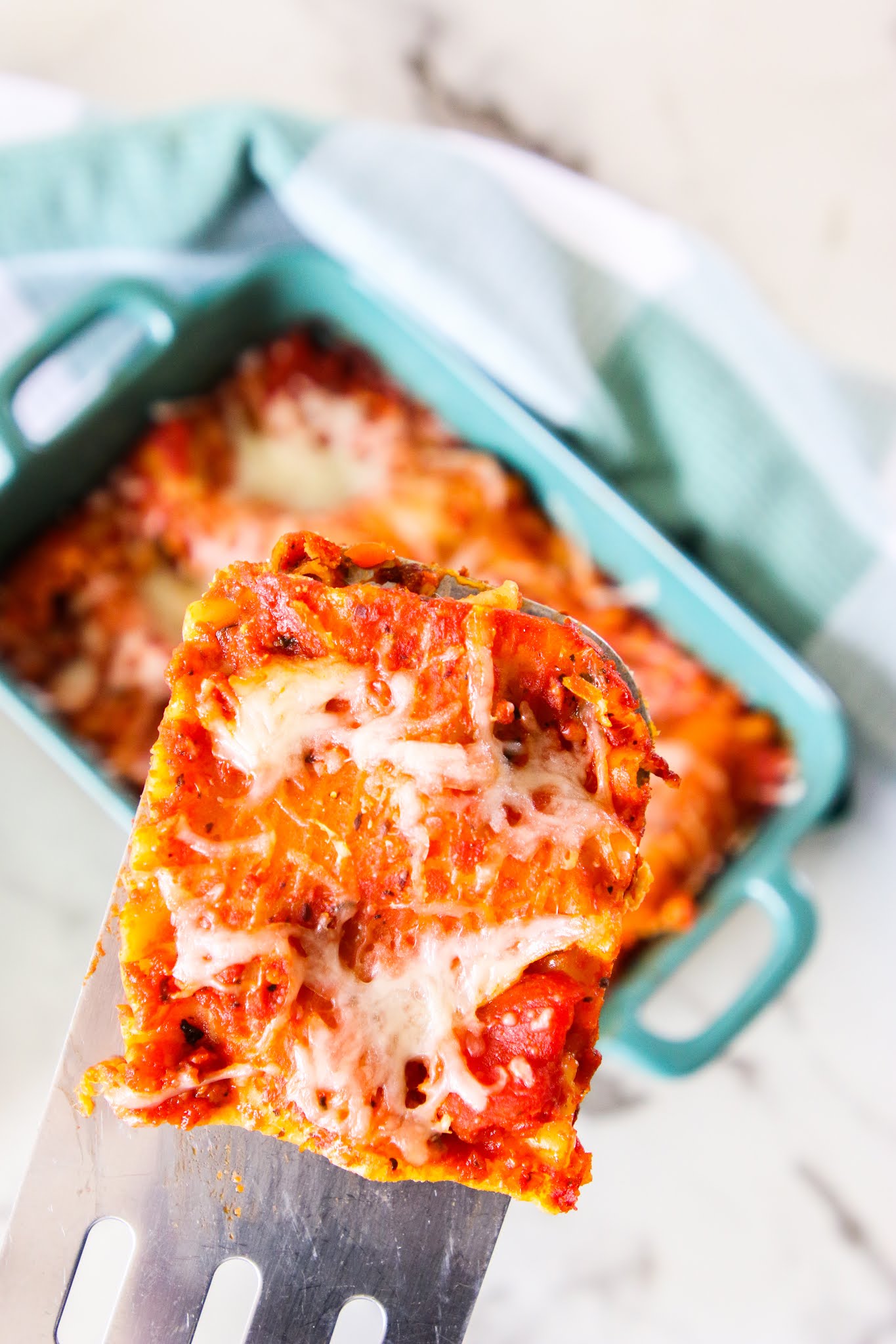 Lasagna in a green baking dish with a green plaid towel in the background on a marble table.