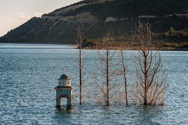 Czubek dzwonnicy zatopionego kościół św. Mikołaja, w Alassie na Cyprze, wystający z kilkoma drzewami nad taflą wody.