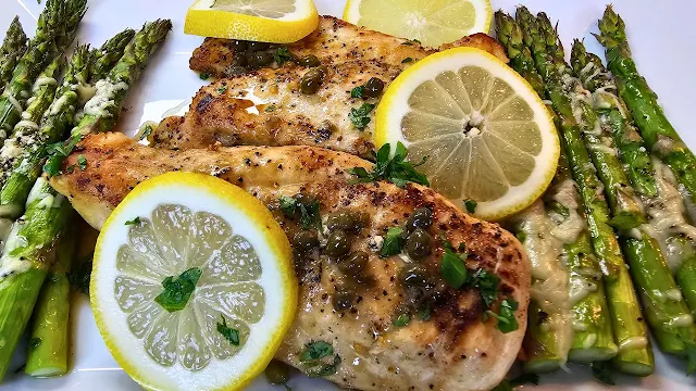 Chicken Piccata on a white platter with lemon slices, and roasted asparagus.