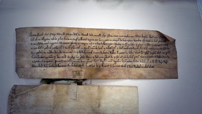 An old document, brown and irregular, with eight lines of dark old fashioned writing, it is attached by the tab for the seal to another document which is turned face down