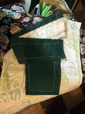 A small pile of not-quite-rectangular piece of fabric, one side dark hunter green and the other the reverse of a teal, white, and green tropical floral, stitched with white about half an inch from the edges, held on the lap of someone wearing a black calaveras-print skirt.