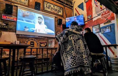 Two fans, one in an LA Dodgers serape, watch the match with a big Great Britain flag on the wall