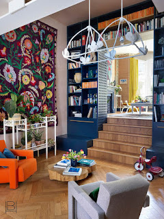 Beata Heuman living room with blue painted library shelves, bold rug hung as art, orange eighties organic modern chair, and live edge wood coffee table