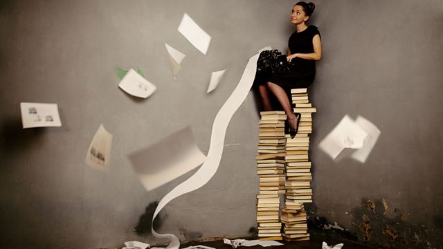 Foto de una chica sentada sobre una montaña enorme de libros mientras llueven hojas escritas