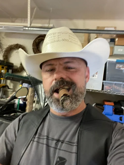 Grey beard, cowboy hat, t-shirt matches facial hair and leather vest smoking a cigar