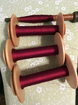 Three wooden spinning wheel bobbins and one long wooden bobbin, all thickly wound with extremely fine deep garnet thread, on a pale-green floral patterned ironing board.