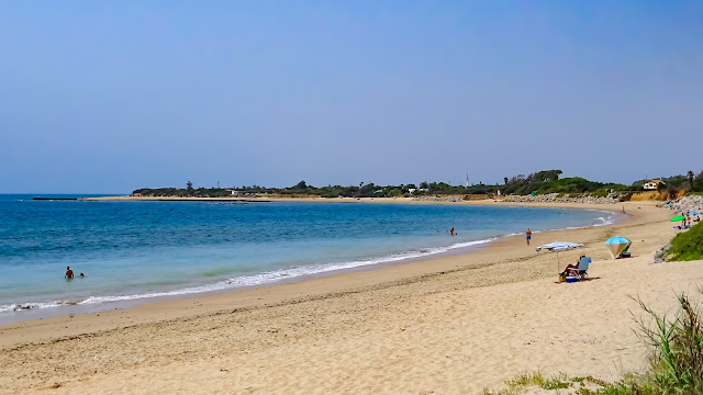 Gran playa llana de arena en forma de herradura y con las azules aguas del mar a su frente.