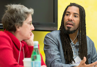 Photo of Kathi Wolfe and L. Lamar Wilson. They sit at a table side by side and Lamar is speaking. Kathi has short gray hair, wears a red sweater and glasses She is listening intently. Lamar has on a grey jacket, a light mustache and well trimmed beard. He is wearing his hair in long locks. There is a yellow wall behind them.