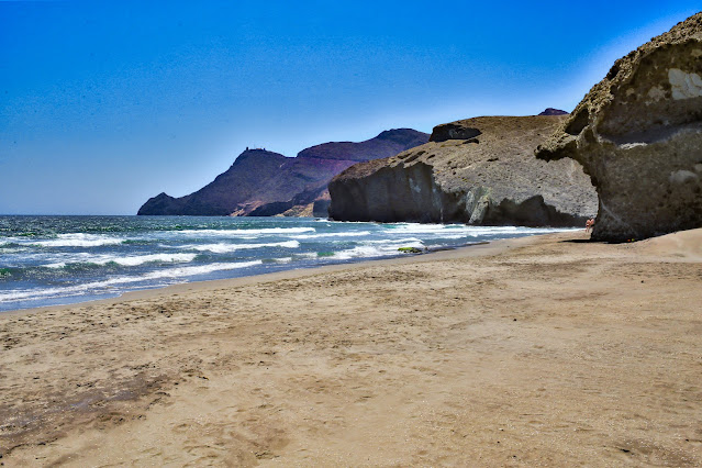 Playa de arena fina con el mar y paisaje rocoso volcánico al fondo