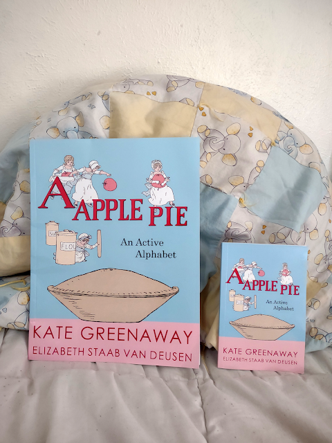 A large 8.5x11 book sits next to a small 4x6 book as a size comparison. Both books are A Apple Pie: An Active Alphabet, by Kate Greenaway and Elizabeth Staab Van Deusen. The cover is blue and pink, with an illustration of a large pie and four small children with baking supplies.